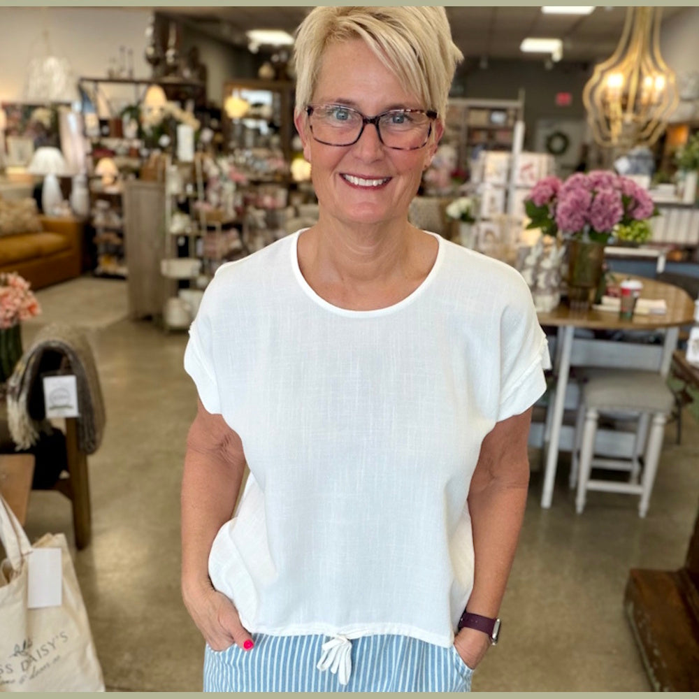A smiling woman with short blonde hair and glasses wears a white linen drawstring top and blue striped pants.
