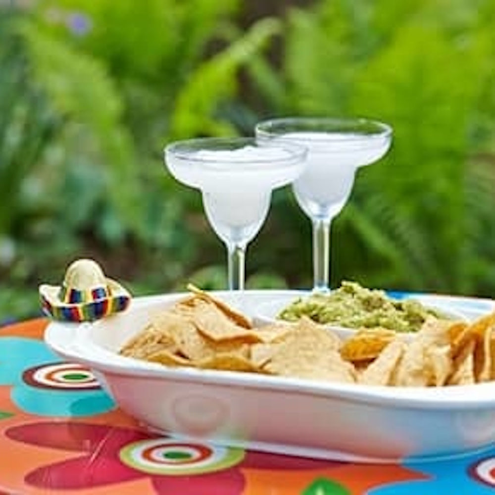 A tray of tortilla chips and guacamole next to two frozen margarita glasses on a colorful outdoor table.