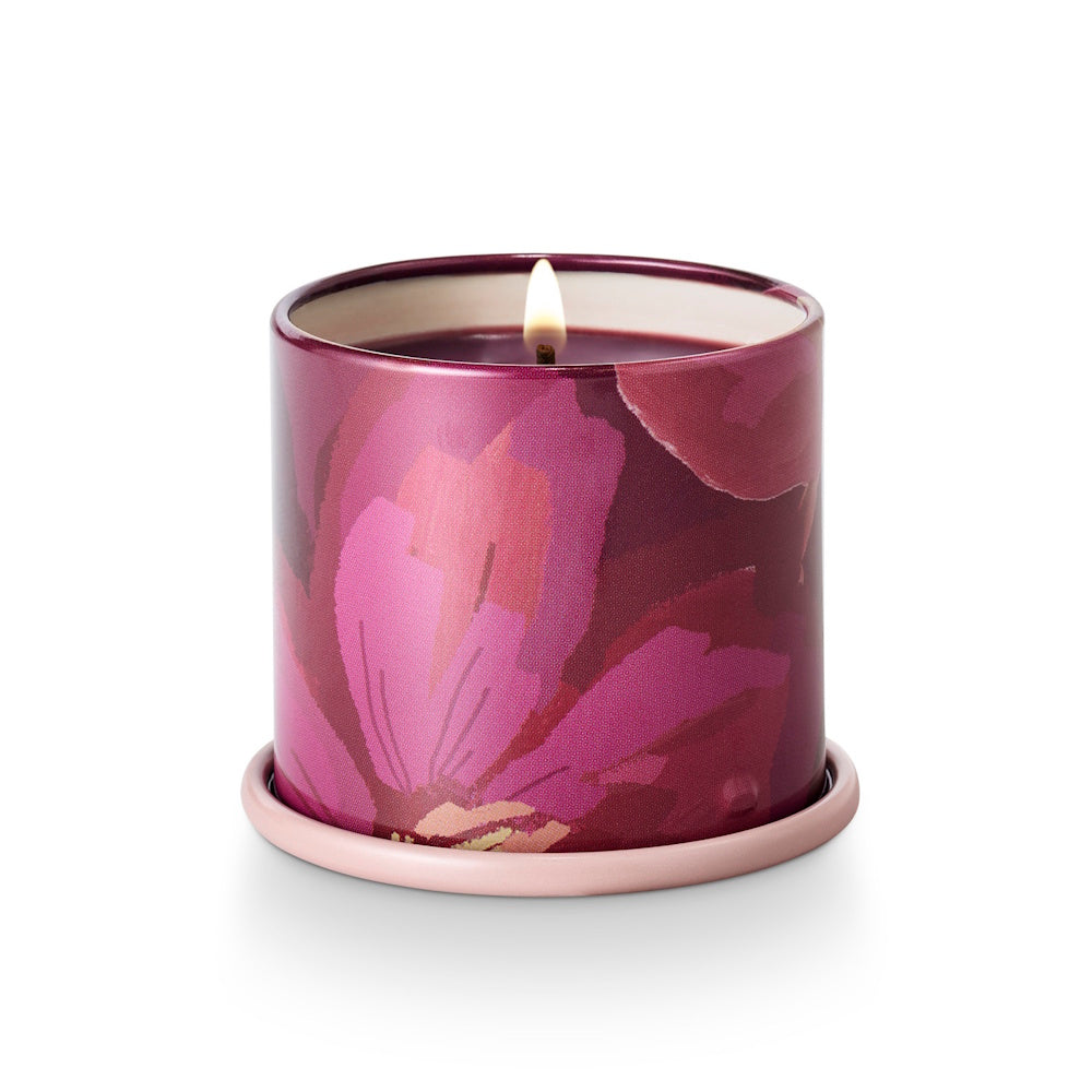 Lit candle in a magenta and pink floral tin container resting on a small pink saucer, white background.