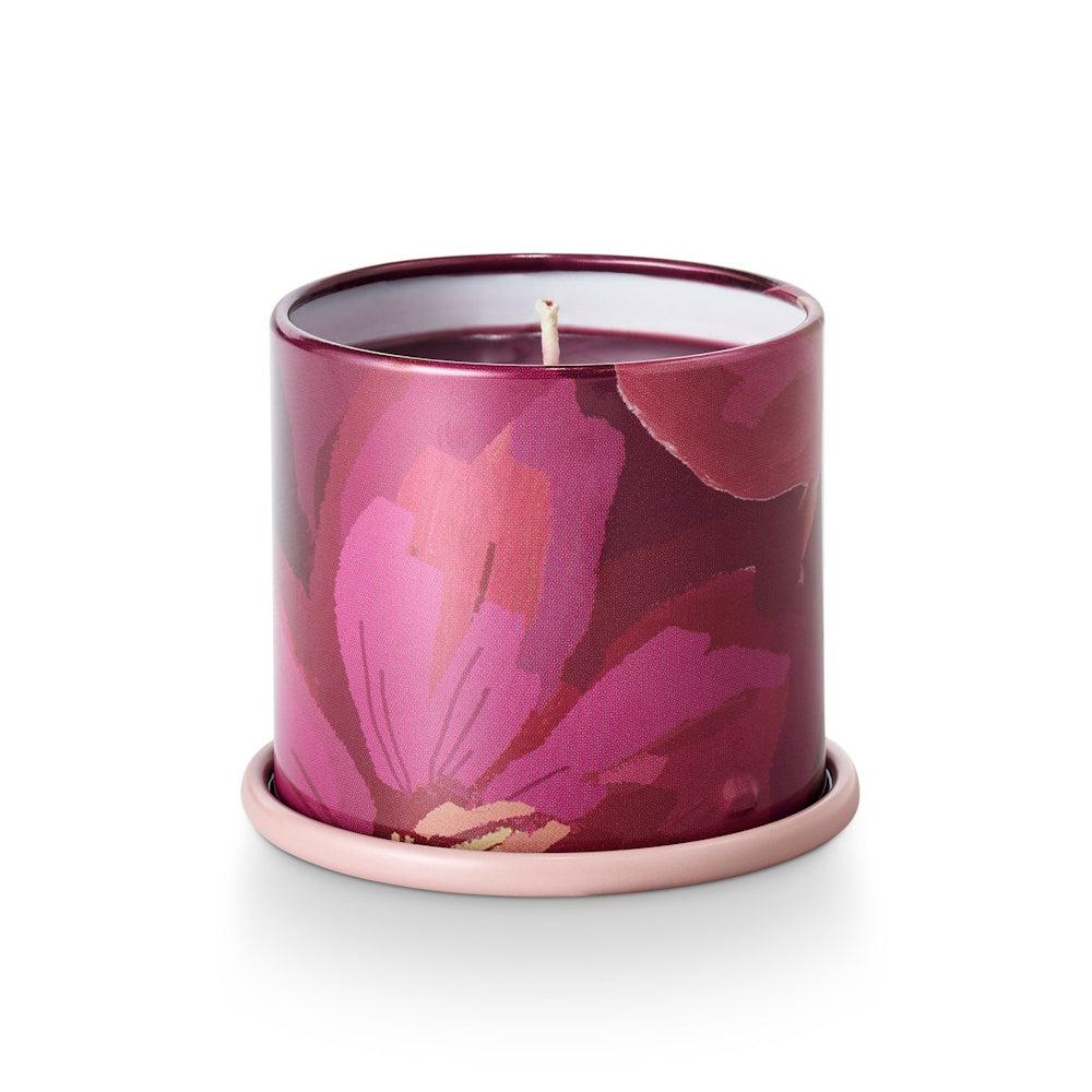 A small candle in a pink and purple floral tin vessel, resting on a matching light pink saucer base.