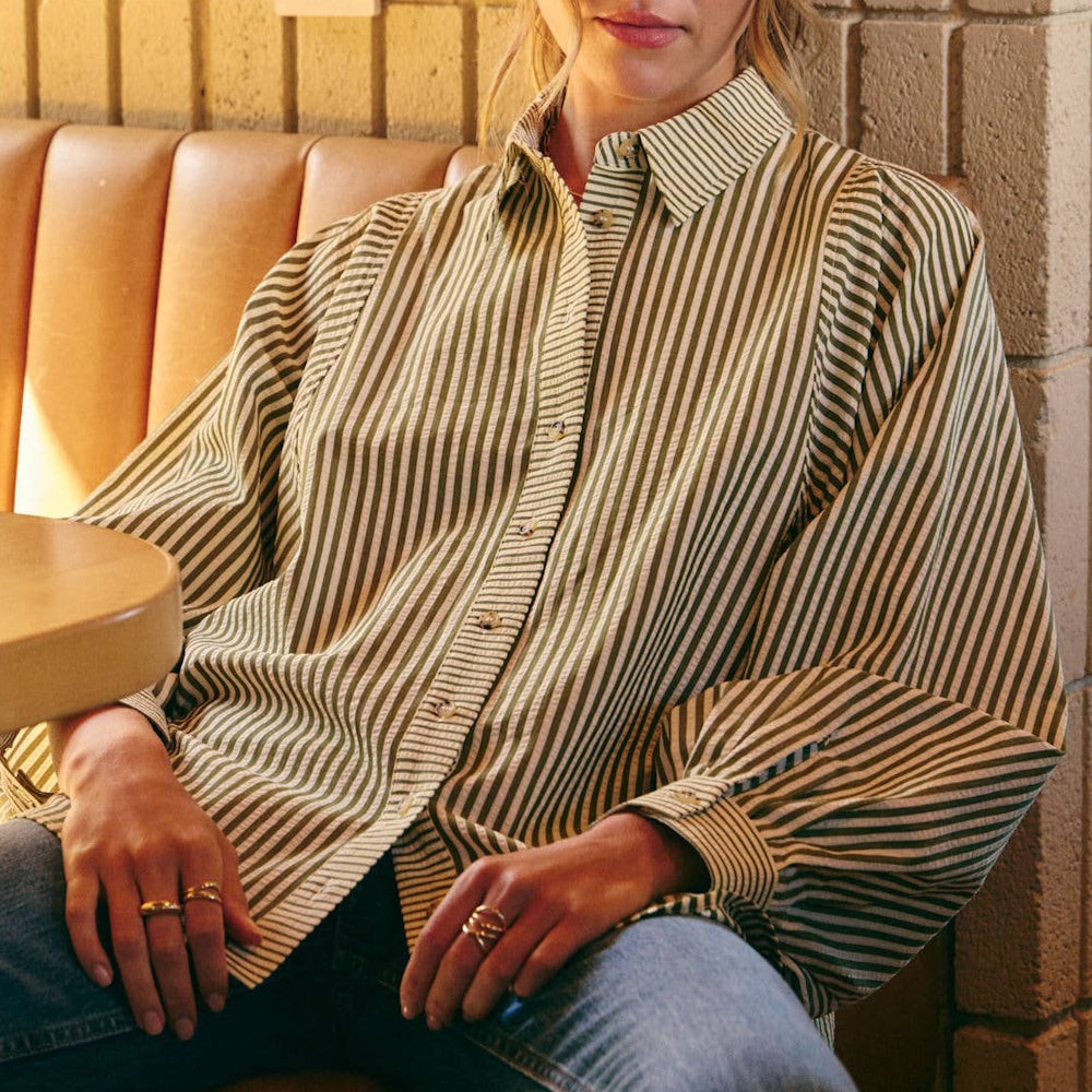 Woman sitting in a cafe booth wearing an olive green and white striped, relaxed-fit button-down shirt.