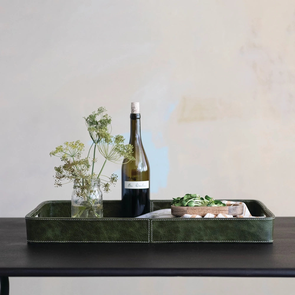 Olive green decorative leather tray with handles, holding a wine bottle, dried herbs, and small snacks on a dark table.