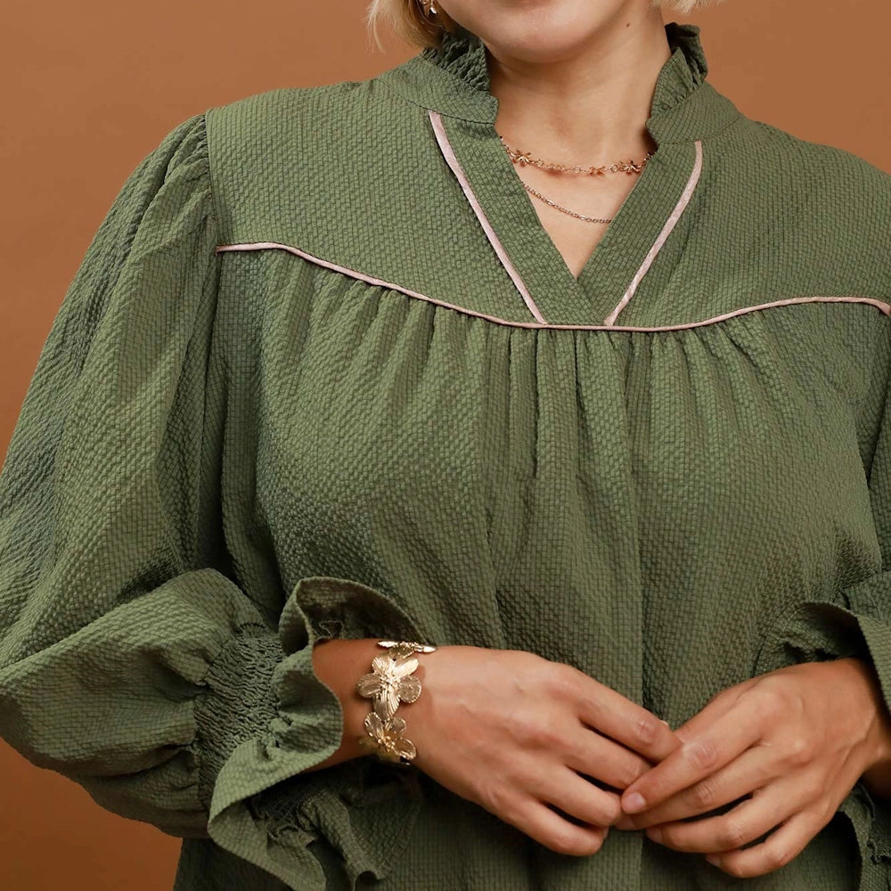 Close-up of a woman in an olive green textured blouse with pink piping detail and ruffled cuffs.