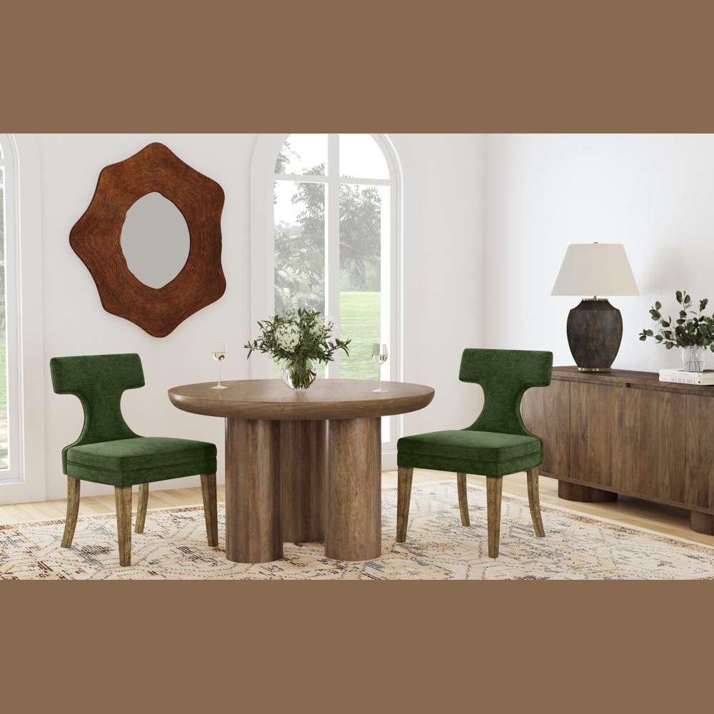 Modern round wood dining table with two green velvet chairs, a unique wall mirror, and a wood sideboard in a bright room.