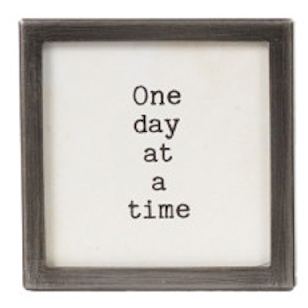 A square silver-framed sign with the text "One day at a time" in black typewriter font.