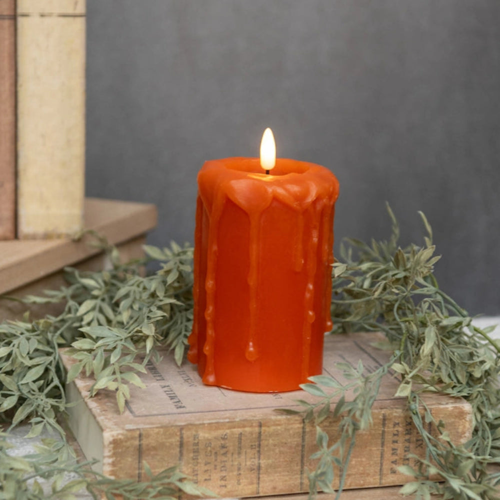 Lit orange pillar candle with wax drips, resting on vintage books with greenery accents.
