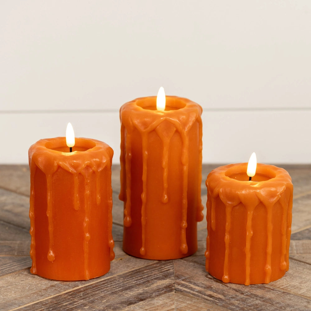 Three glowing orange flameless pillar candles with wax drips resting on a wood surface.