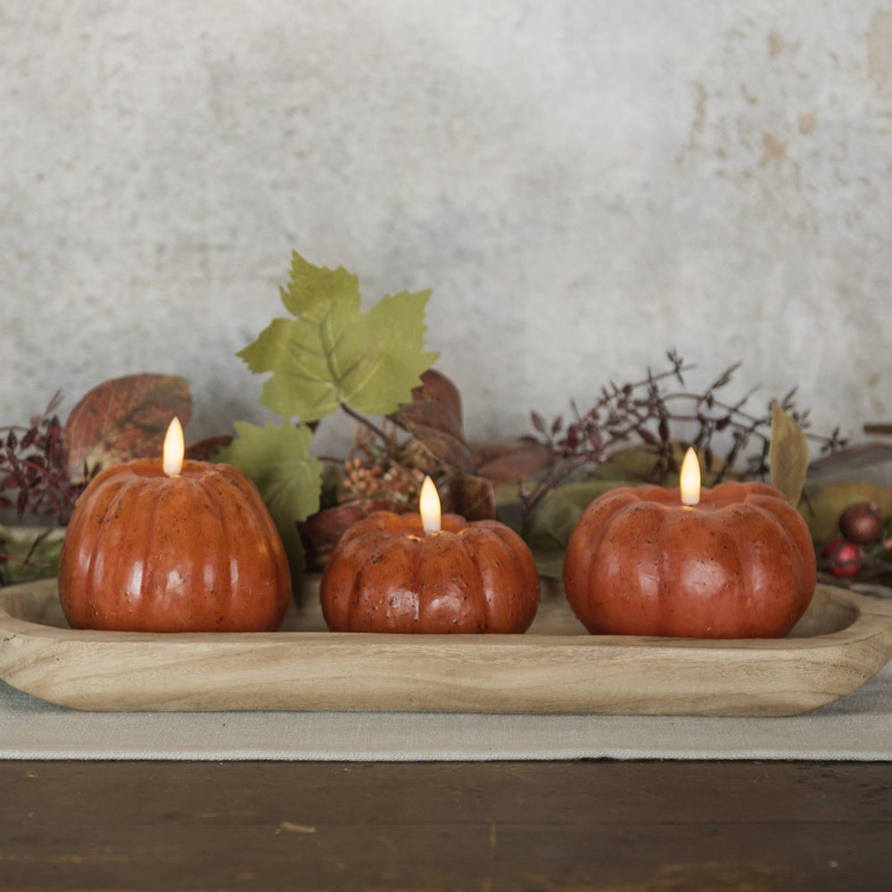 Three orange pumpkin-shaped flameless candles lit in a wooden tray with autumnal leaves.
