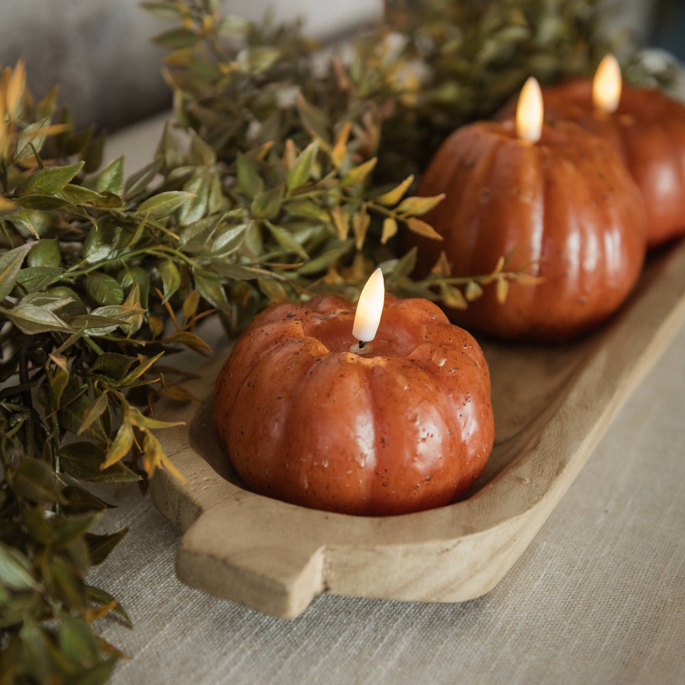 Three lit orange pumpkin-shaped candles in a wooden dough bowl with leafy greenery.