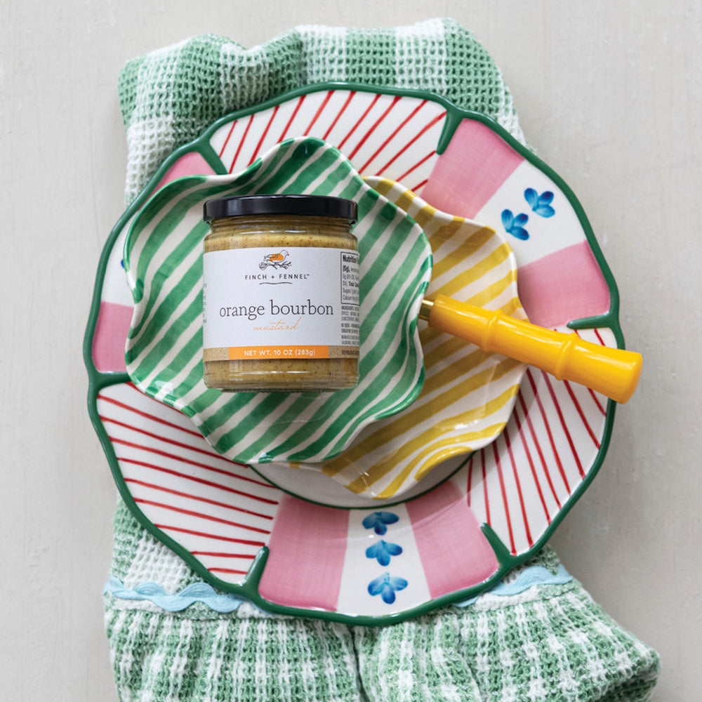 A jar of Finch + Fennel orange bourbon mustard sitting on top of a stack of colorful, patterned plates.