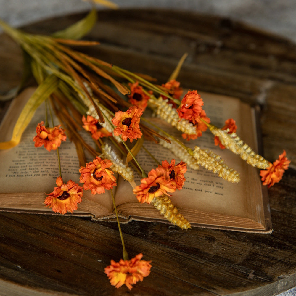 A bouquet of orange flowers and artificial wild grass resting across an open antique book on a wooden surface.
