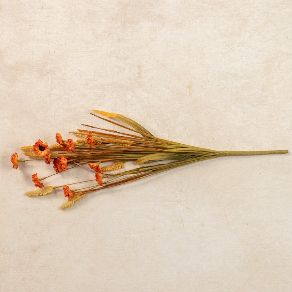 An artificial spray of orange wildflowers, grasses, and wheat sprigs lying against a textured beige background.