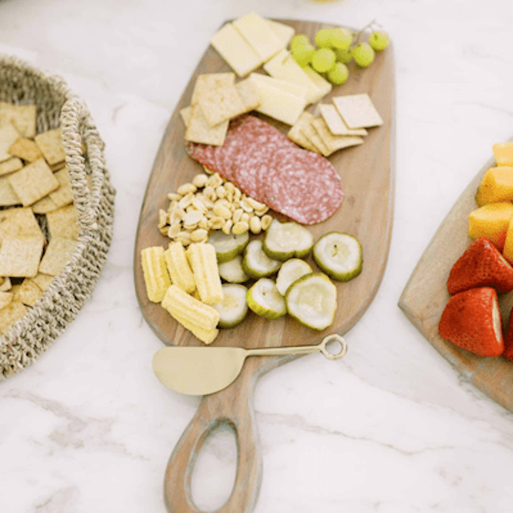 A charcuterie board with crackers, cheese, grapes, salami, peanuts, pickles, and baby corn next to a knife.