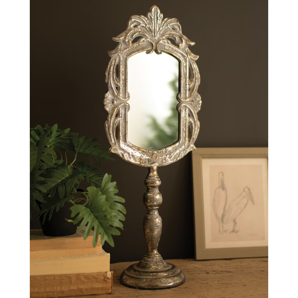 Antiqued metal pedestal mirror with an ornate frame set on a table beside books and artwork.
