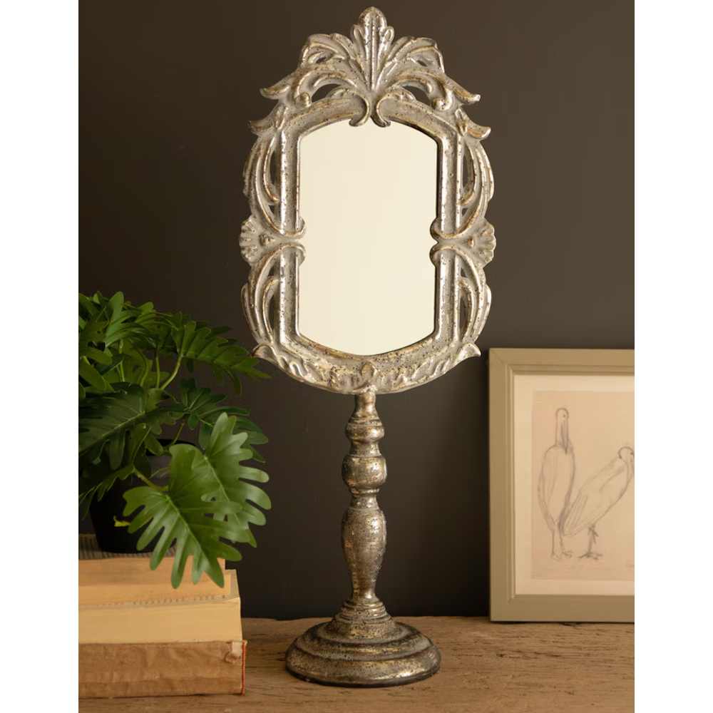 Vintage ornate metal vanity mirror on a pedestal stand set against a dark wall and wooden desk.