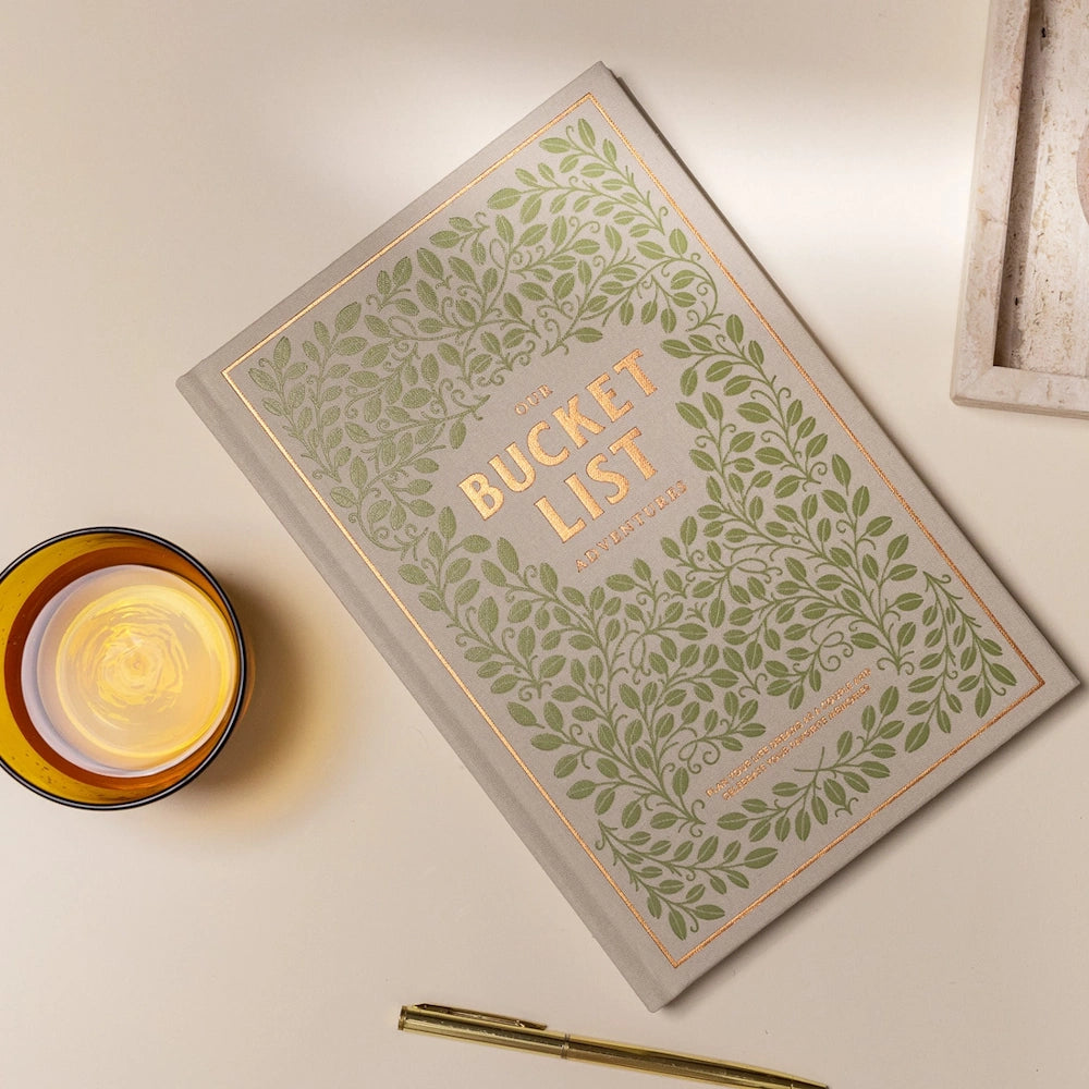 Our Bucket List Adventures journal with green leaf patterns, gold foil text, a pen, and a drink on a neutral surface.