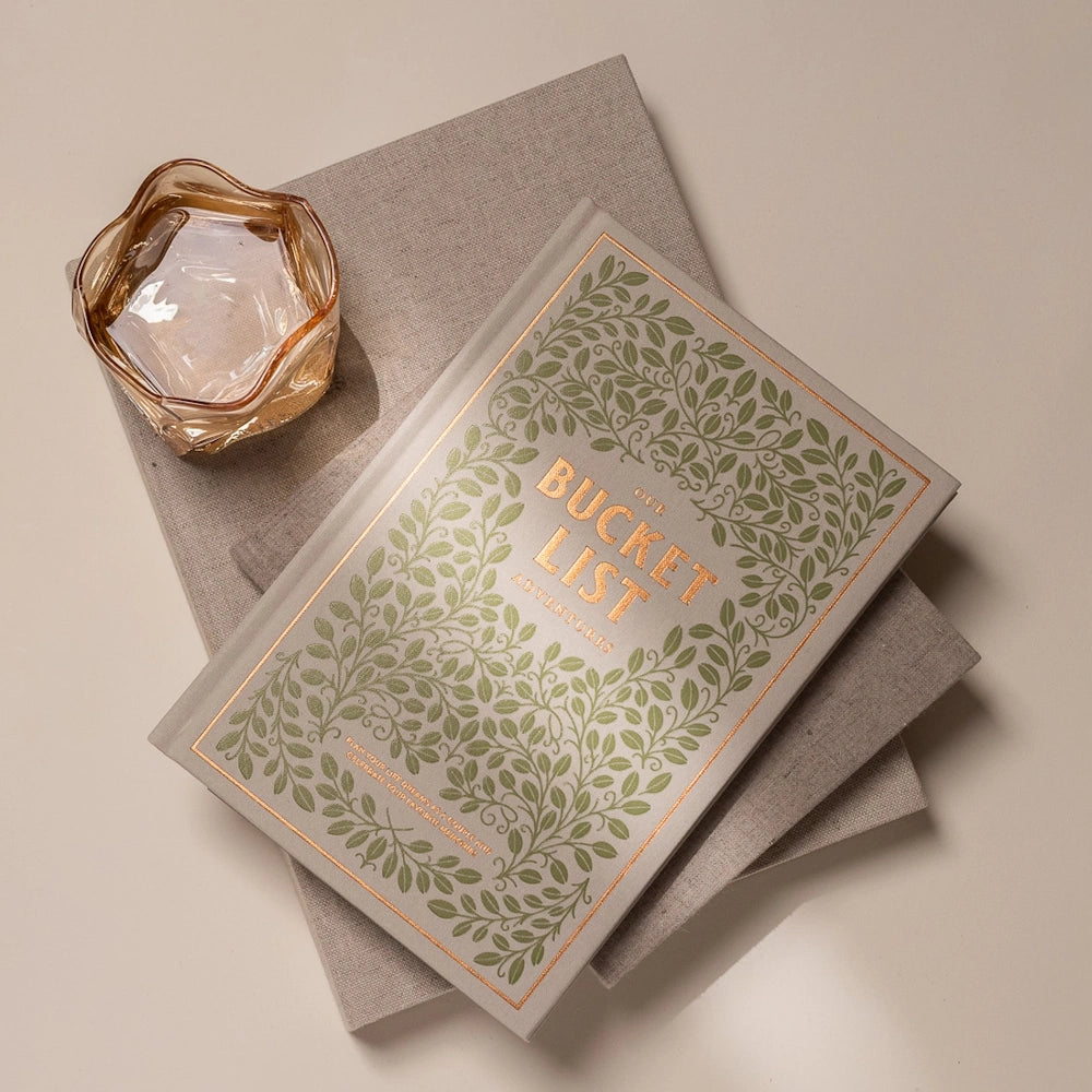 Our Bucket List Adventures journal with a gold-foiled leaf design, resting on linen books next to a decorative glass.