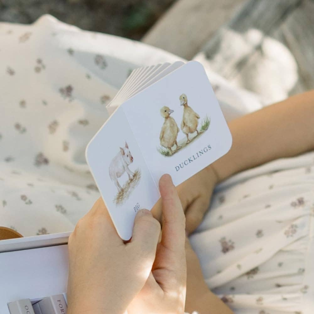 Child holding an open Our Very Little Library board book featuring watercolor illustrations of a pig and ducklings.