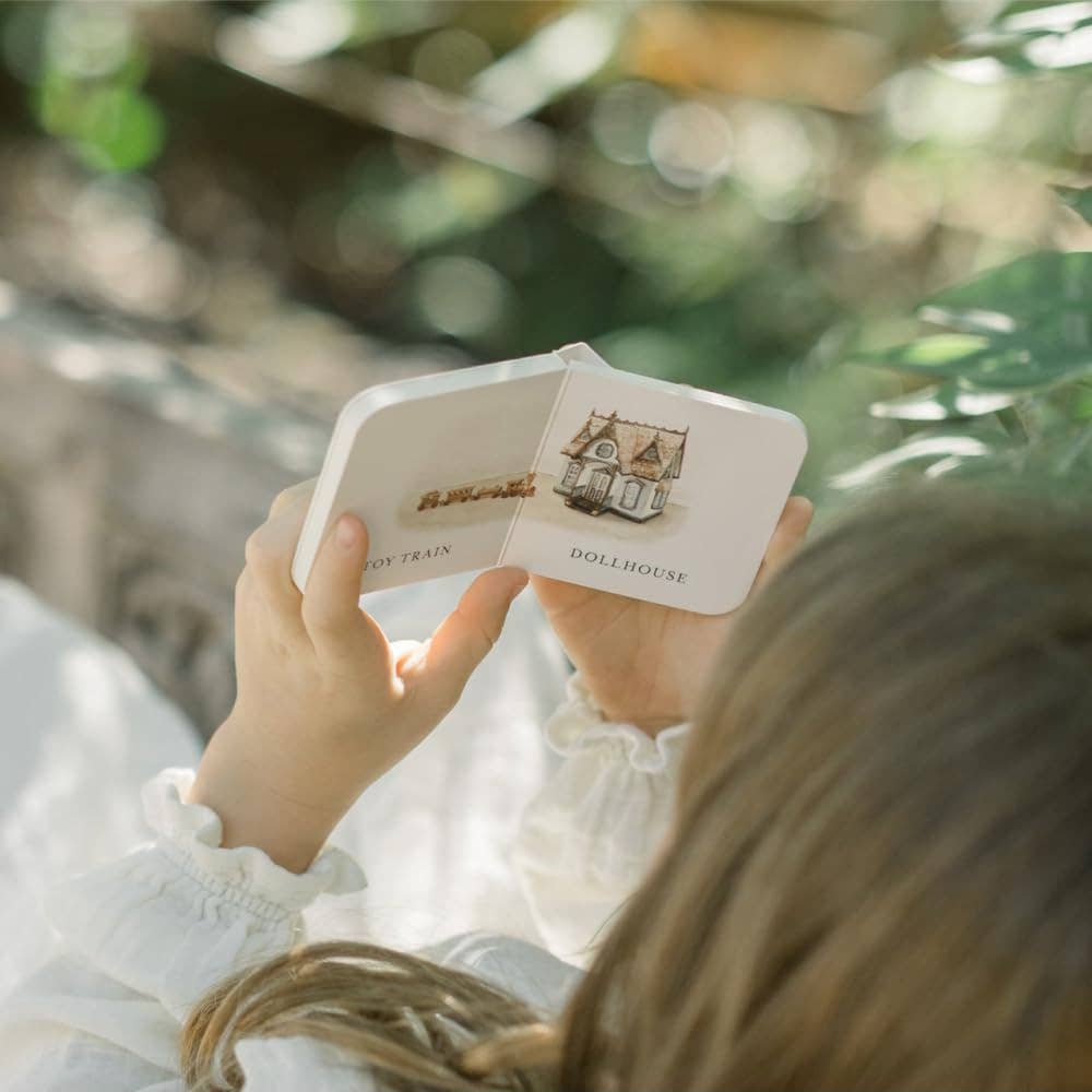Child reading Our Very Little Library board book featuring toy train and dollhouse illustrations in a natural setting.