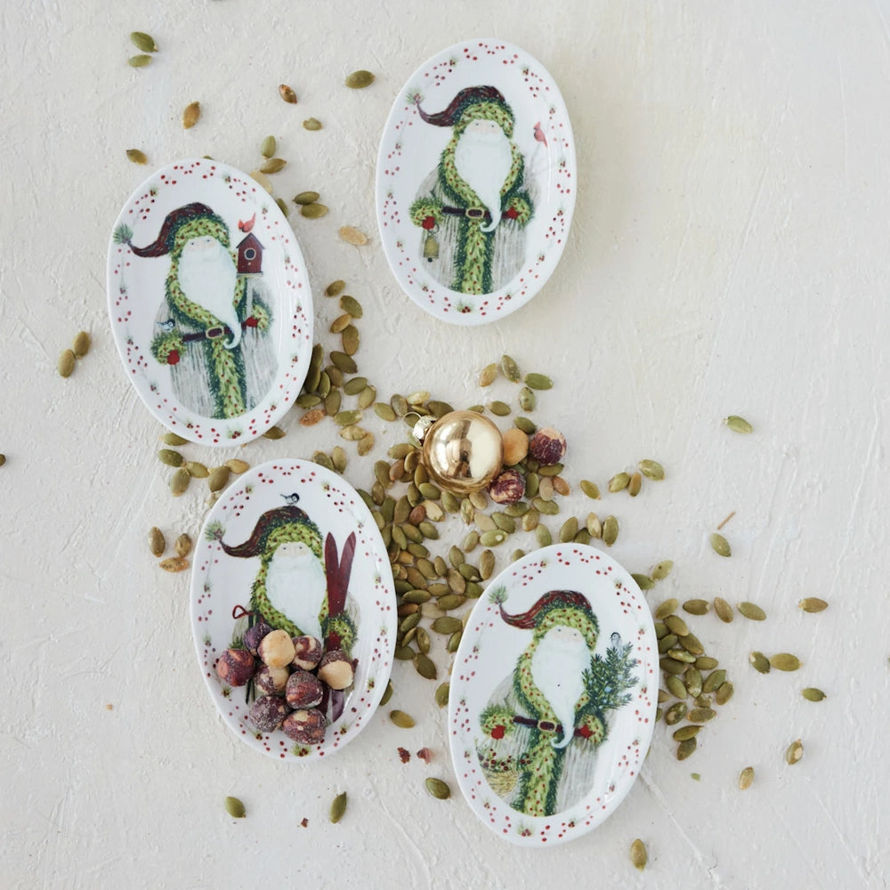 Four oval Santa platters arranged on a textured surface surrounded by seeds, nuts, and a gold ornament.