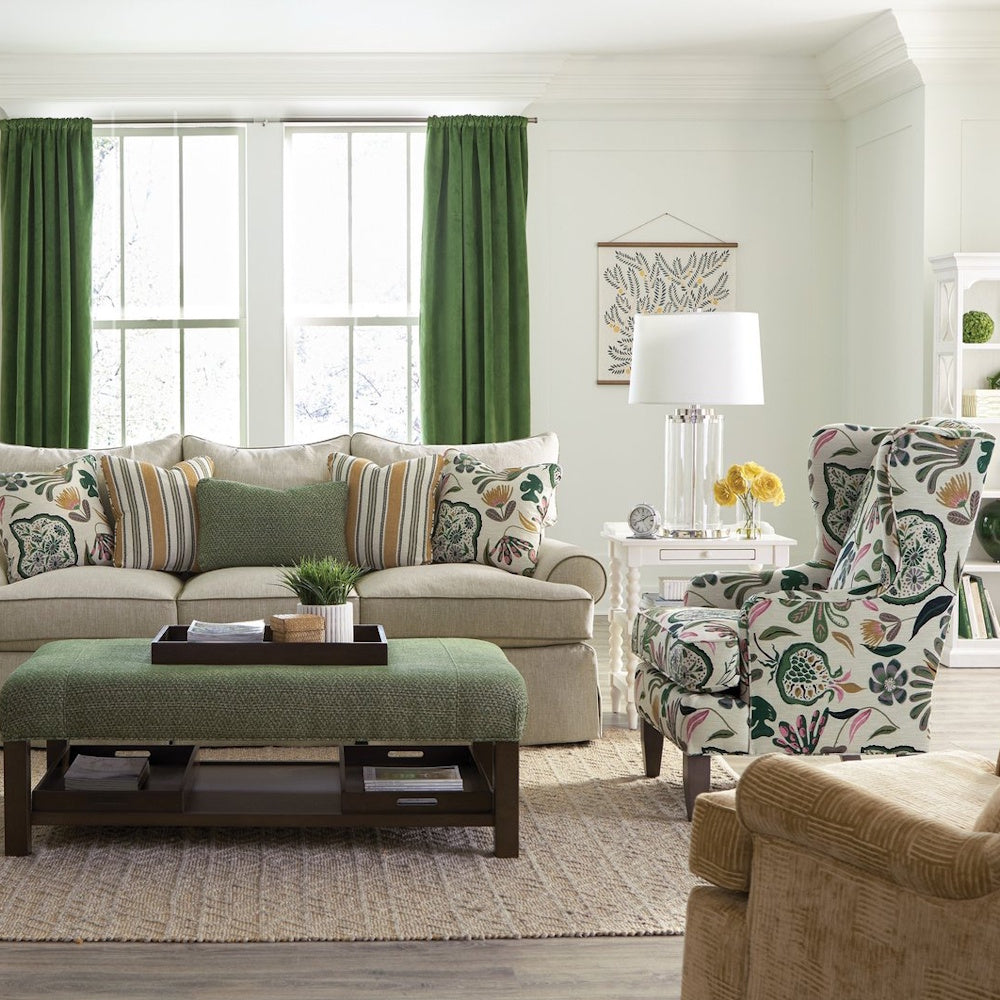 Floral wingback armchair in a bright living room beside a sofa, green ottoman, and large windows.