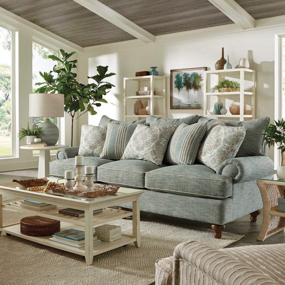 Light blue sofa with patterned pillows in a bright living room with white tables and house plants.
