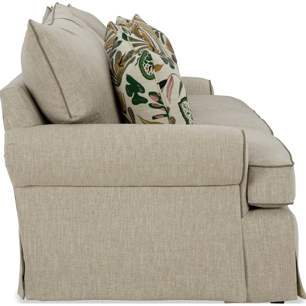 Side profile of the beige skirted three-cushion sofa with patterned accent pillows on a white background.
