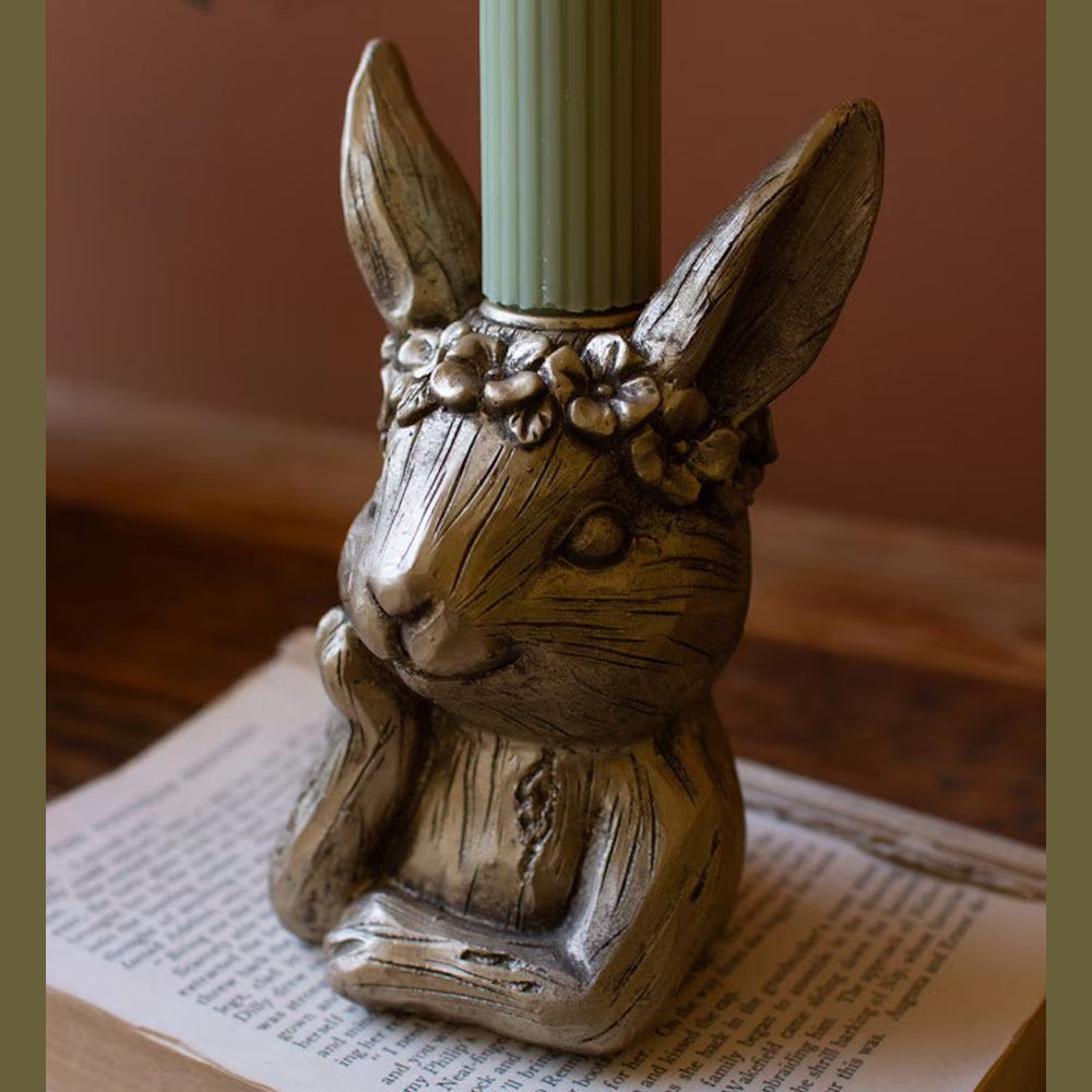 Close-up of a silver resin rabbit taper candle holder with a flower crown, resting on an open book with a green candle.