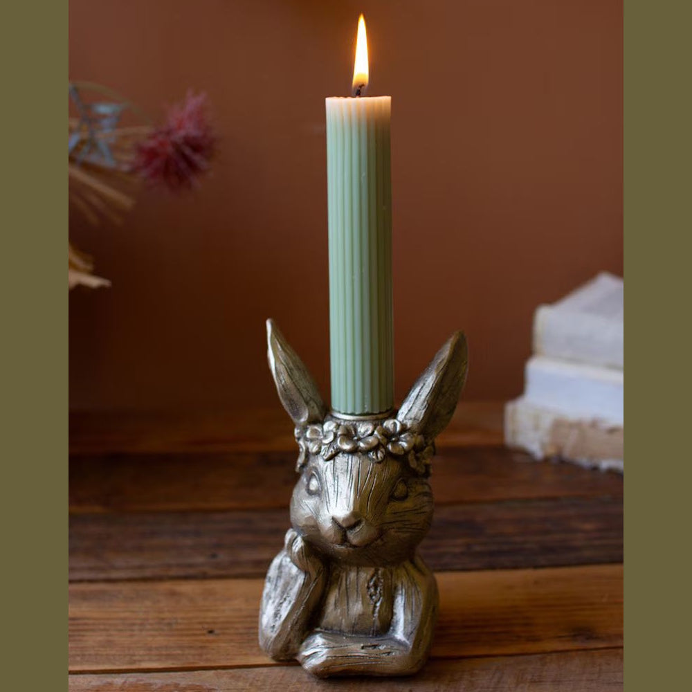 Silver painted resin rabbit taper candle holder with a floral crown holding a lit green ribbed candle on wood surface.