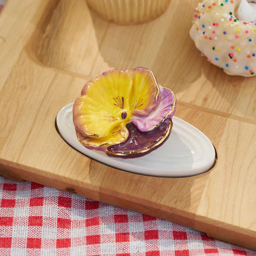 Yellow and purple ceramic pansy flower on a white base set in a wooden tray with pastries.