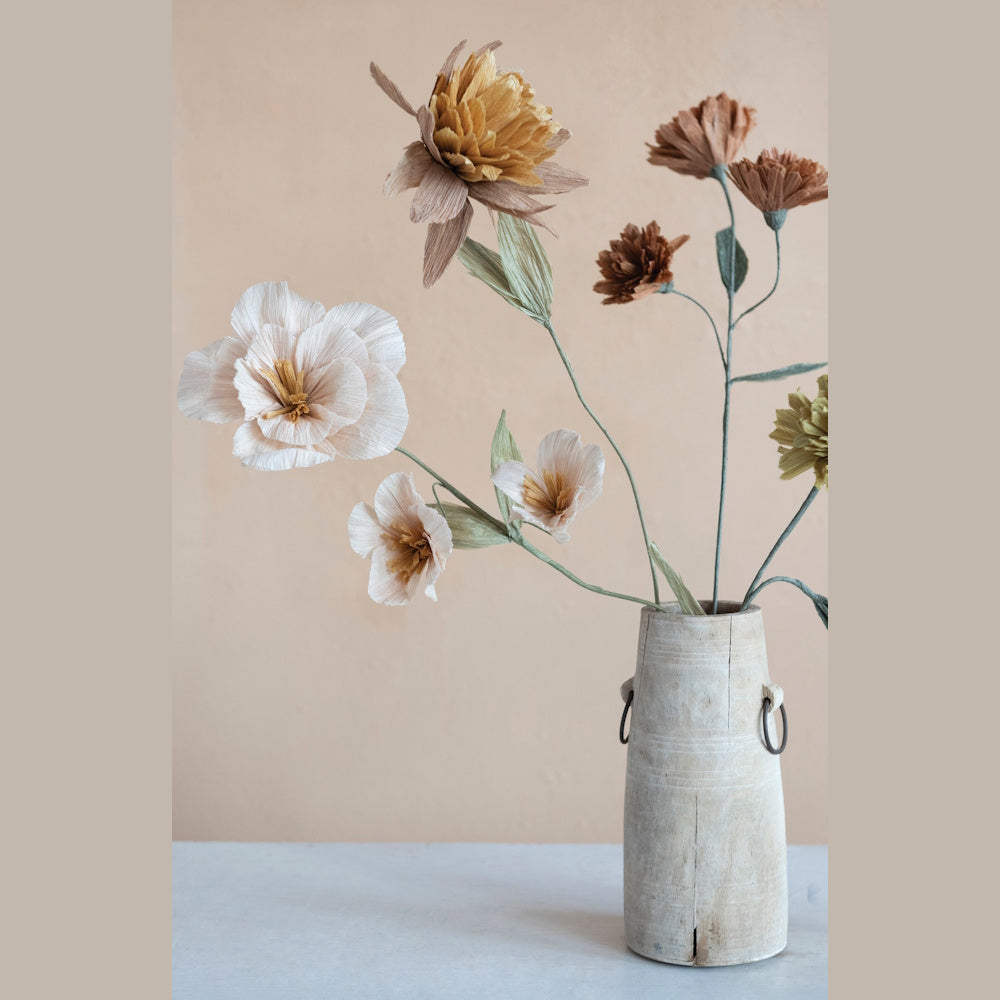 Assorted crepe paper flowers in muted neutral tones arranged in a rustic wooden vase against a peach wall.