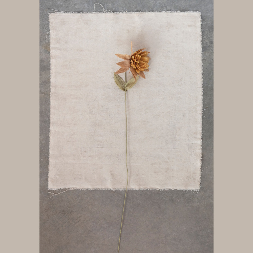 A single long-stemmed, golden-brown paper flower on a frayed beige linen cloth.