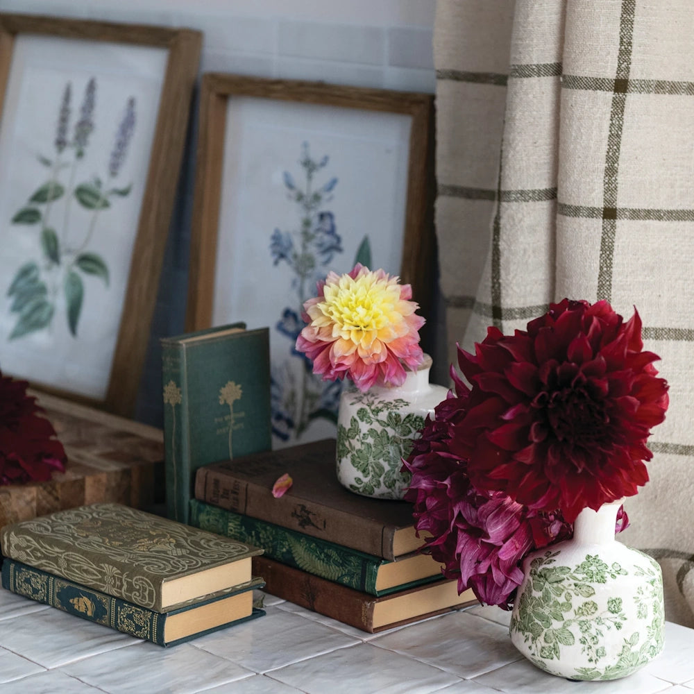 Vintage books and dahlias in floral vases on a tiled surface with framed botanical art in the background.