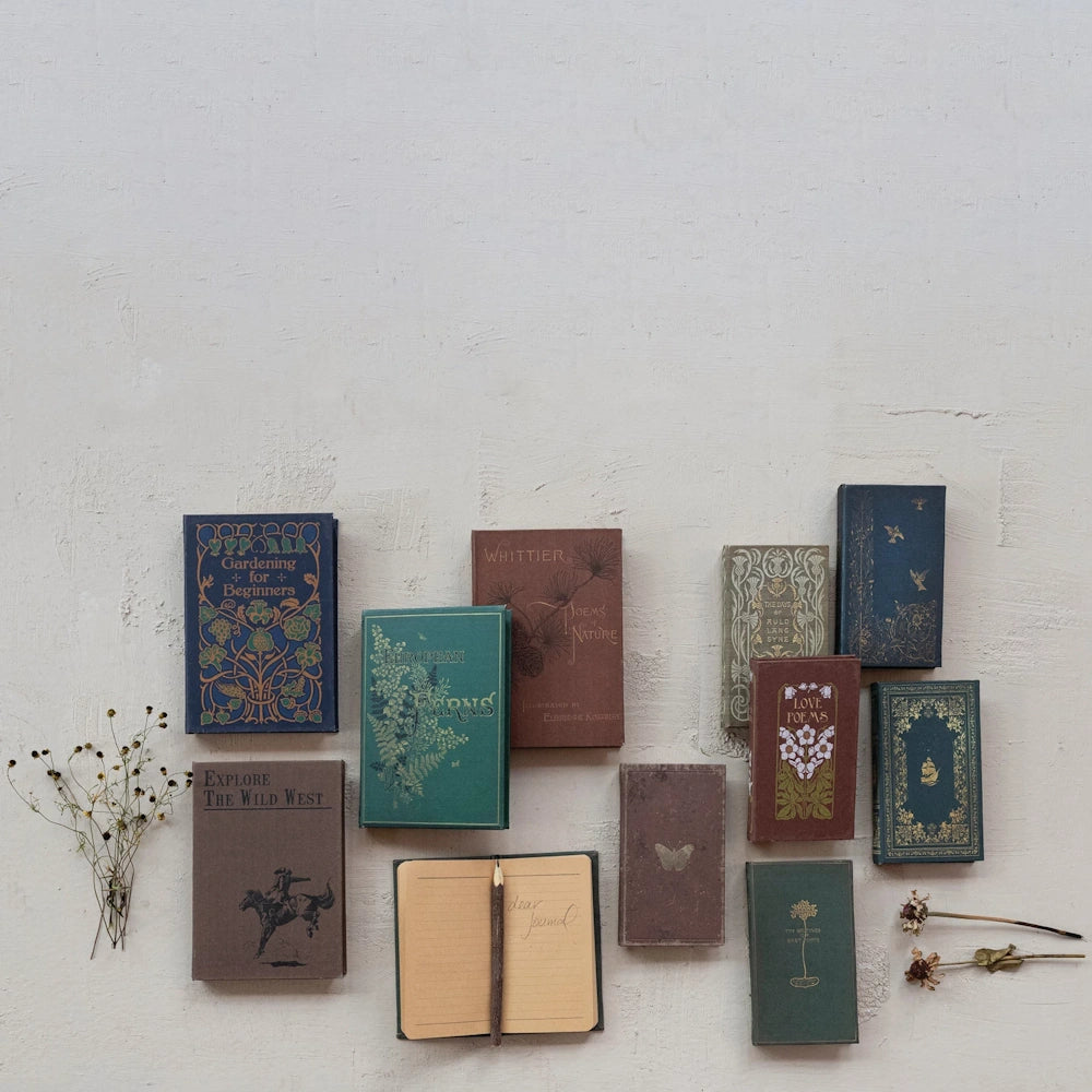 Vintage-style notebooks and books with floral and nature designs arranged on a white wall with an open journal and pencil.