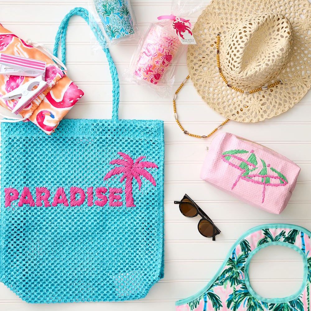 Flat lay of beach essentials featuring a blue mesh tote with pink "PARADISE" embroidery and a palm tree.