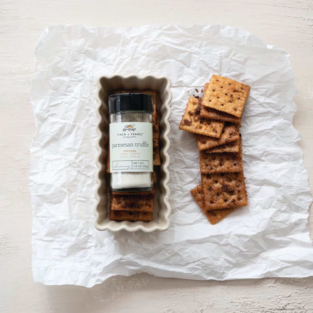 A Finch + Fennel Parmesan Truffle seasoning shaker sits on a tray of square crackers on white paper.