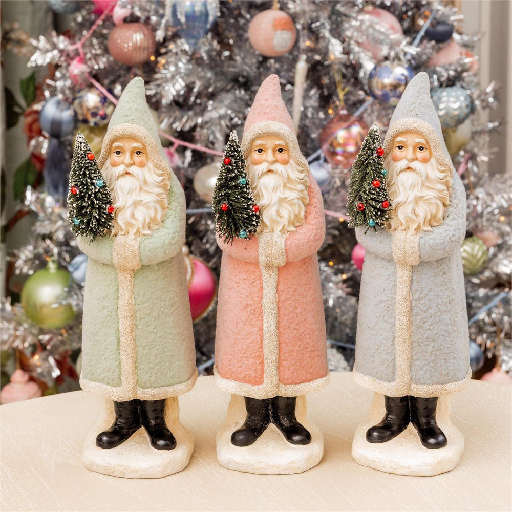 Three pastel Santa figurines holding small Christmas trees in front of a decorated tree.