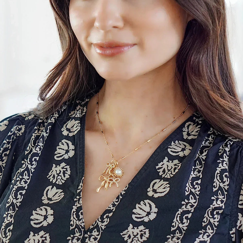 Woman wearing a gold charm necklace featuring a crystal pavé looped bow charm and a pearl pendant.