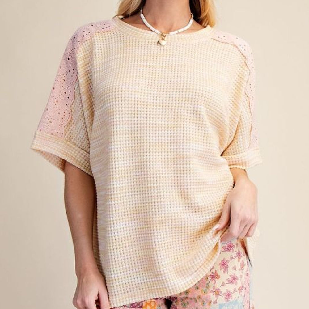 A woman wearing a peach and lemon waffle knit top with pink eyelet lace shoulder detailing and a scalloped trim.