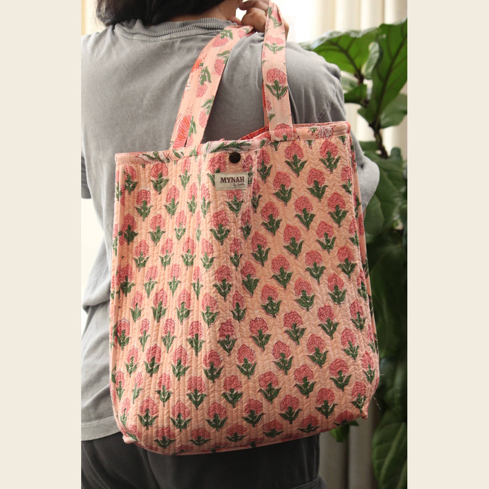 Quilted peach reversible tote bag with pink poppy floral block print and Mynah brand label, carried over a person's shoulder.