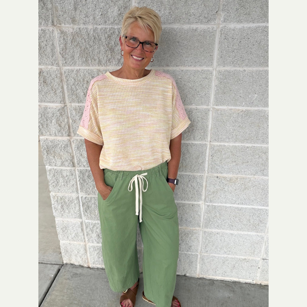 Woman in peach and lemon eyelet lace top and green drawstring pants against a grey brick wall.