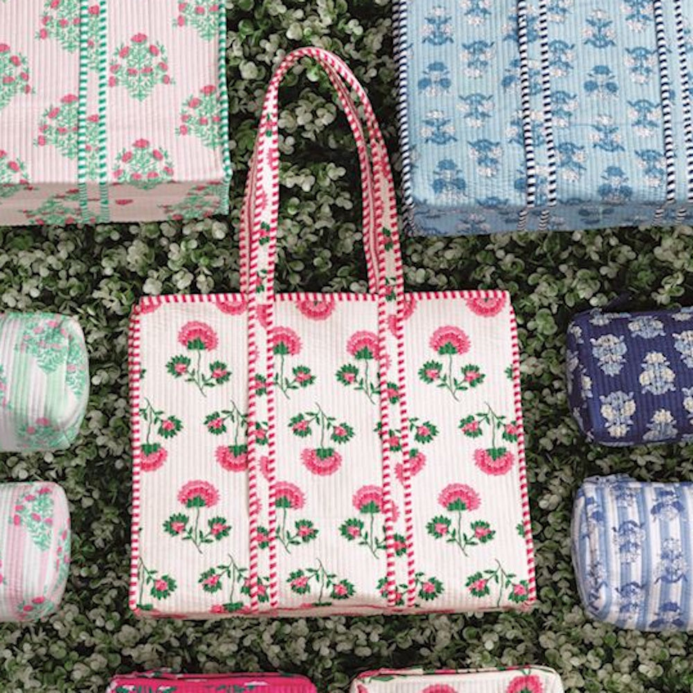 White quilted block-print tote with pink floral motifs and striped trim, surrounded by matching pouches on green foliage.