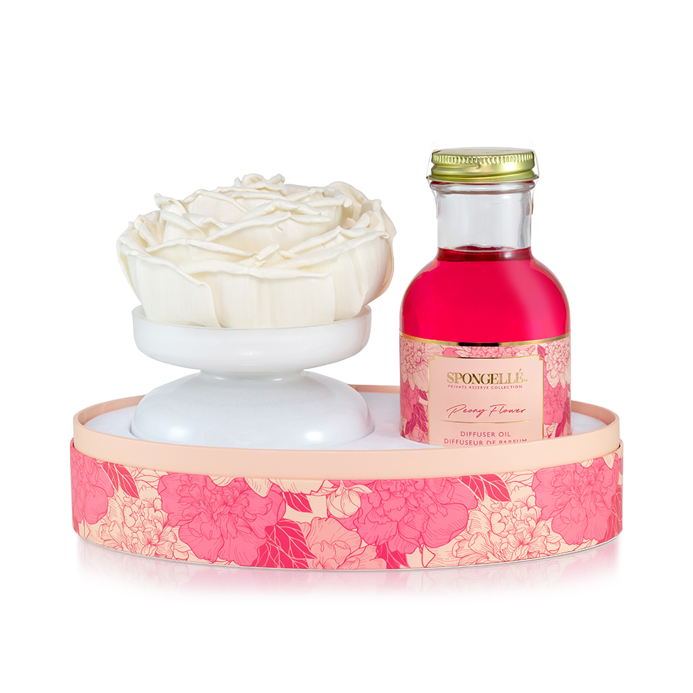 A white ceramic flower diffuser alongside a bottle of vibrant pink diffuser oil in a floral-patterned tray.