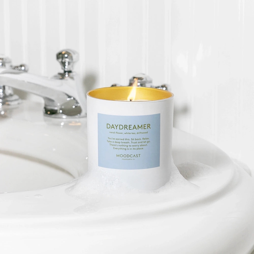 Lit Moodcast Daydreamer candle with a light blue label sitting in soap bubbles on the edge of a white sink.