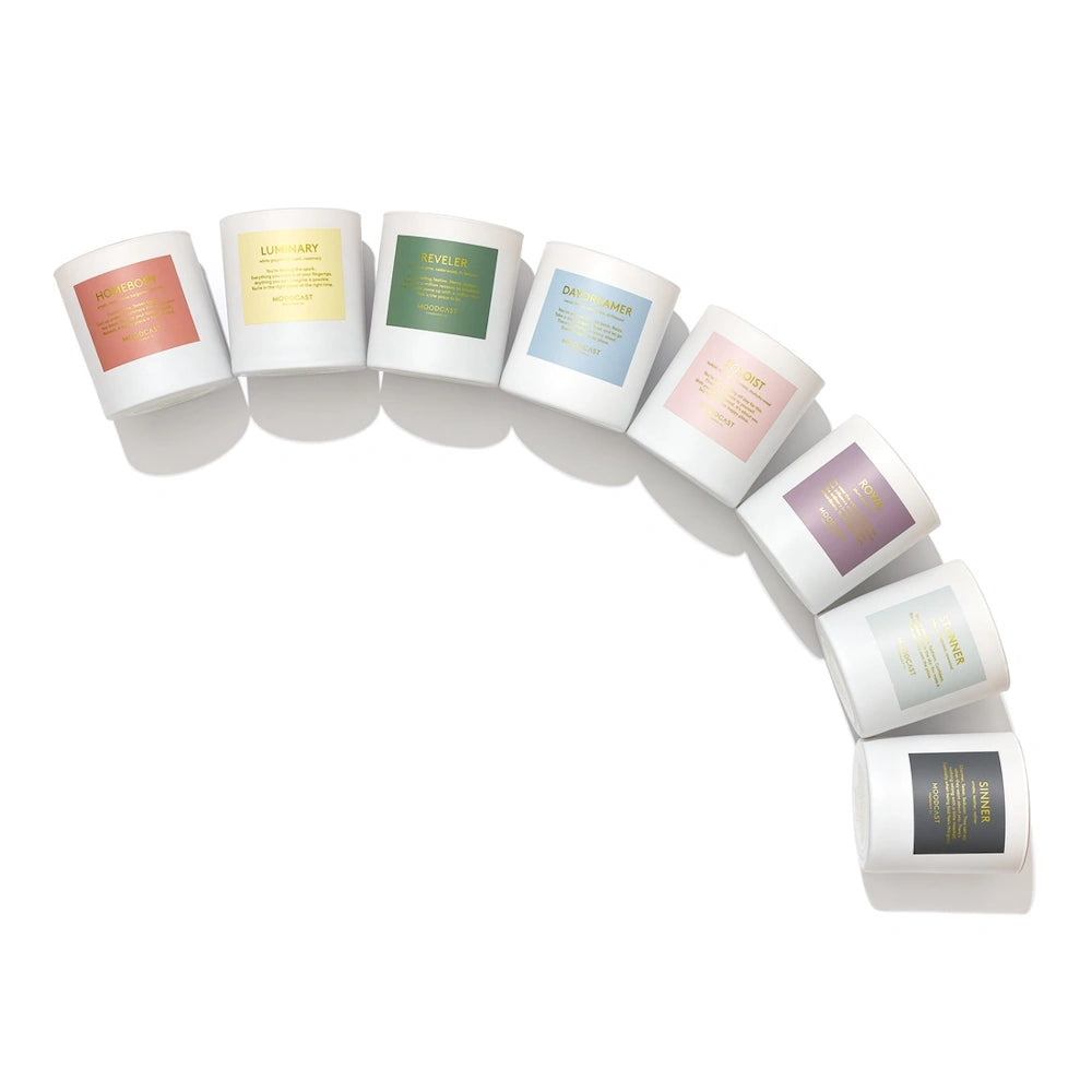 Eight white candles in jar containers arranged in a curve, each featuring a uniquely colored rectangular label.