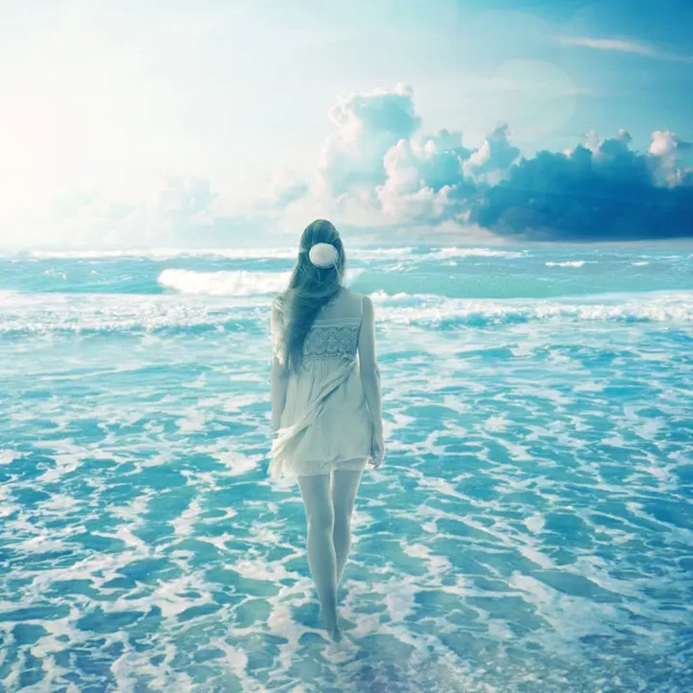 Woman in a white dress walking into bright turquoise ocean water under a dreamy sky with soft clouds.