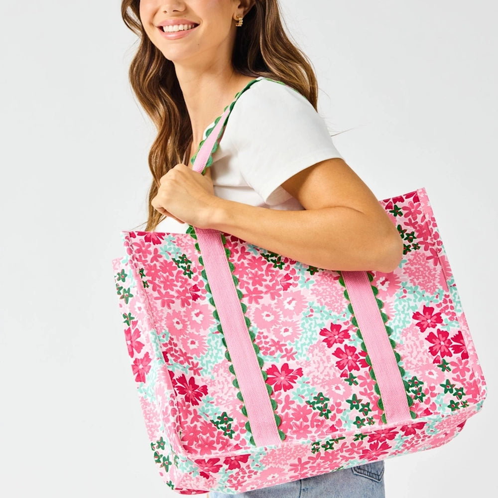 A woman carries a large, pink floral tote bag with green scalloped trim and pink straps over her shoulder.