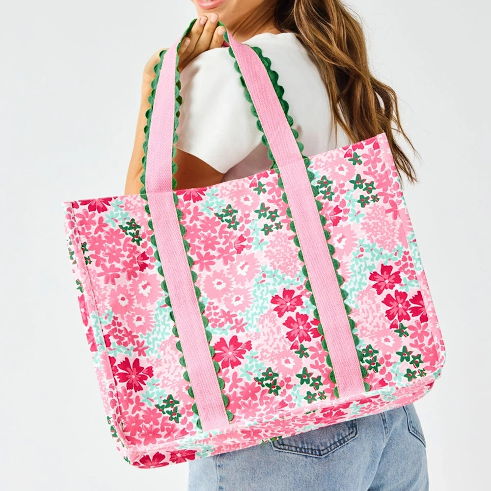 A person carrying a large, floral-patterned pink tote bag with green scalloped trim on its straps over their shoulder.