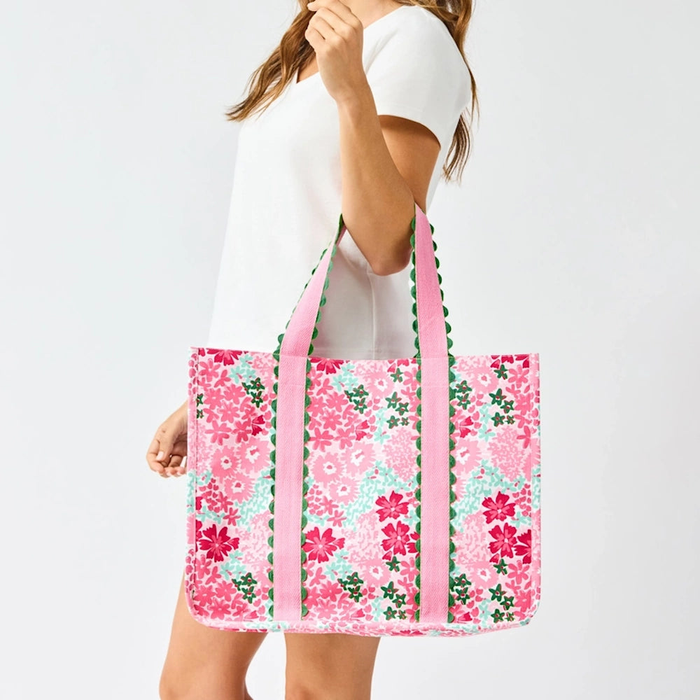 A person holding a pink floral tote bag with pink straps and green scalloped trim against a white background.