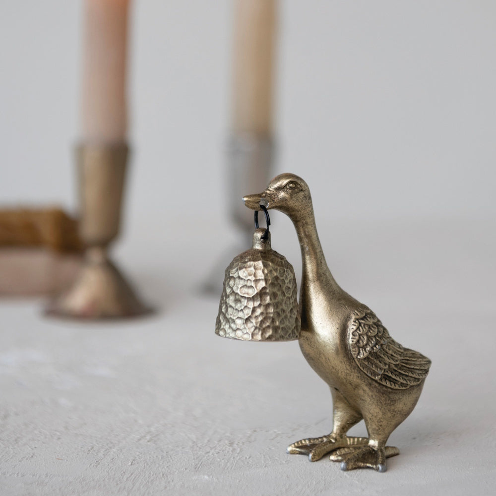 A brass duck-shaped candle snuffer holding a textured bell in its beak, with blurred candles in the background.